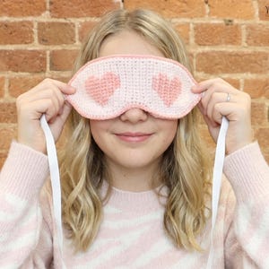 Puede incluir: Un antifaz de crochet con una base de color rosa claro y dos patrones de corazones en rosa más oscuro. El antifaz tiene un borde tejido en rosa claro e incluye una cinta elástica blanca a cada lado para sujetarlo alrededor de la cabeza.