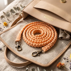 Puede incluir: Un bolso de cuero beige con cierre magnético dorado y el texto ARTISAN en relieve. Dentro del bolso descansa una correa de macramé color melocotón enrollada, con dos mosquetones giratorios de latón envejecido en cada extremo del cordón.