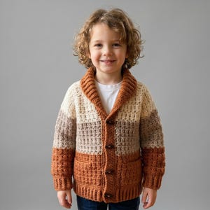 May include: A handmade crochet cardigan in a color block design. The sweater features a shawl collar and is buttoned up. The color scheme includes shades of cream, brown, and rust orange. The cardigan has two front pockets.