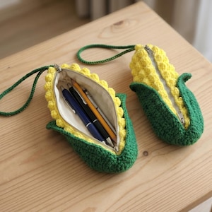 May include: Two corn-shaped crochet pouches with yellow kernels and green husks. One pouch is open, revealing three pens inside, while the other is closed. Both feature a green crocheted wrist strap and a silver metal zip closure mechanism.