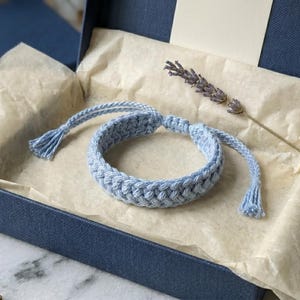 May include: A light blue braided cord bracelet with adjustable sliding knots and tassel ends, presented inside a dark blue gift box lined with beige tissue paper, accompanied by a single dried sprig of lavender resting on the paper.