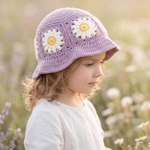 May include: A lavender crochet bucket hat featuring white and yellow daisy flower motifs. The hat is constructed from multiple hexagonal panels, each centered with a floral design, and includes a short, downward-sloping brim around the edge.