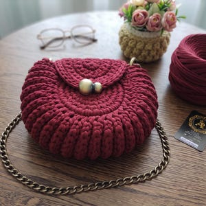 Puede incluir: Un bolso de mano redondo de ganchillo en color rojo oscuro con un patrón acanalado texturizado, un cierre de solapa asegurado con una cuenta metálica grande de dos tonos y una correa de cadena de metal color oro antiguo.