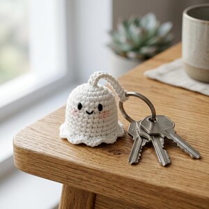 May include: A white crocheted ghost keychain featuring a small bell-shaped body with embroidered black eyes, a black mouth, and soft pink cheeks. The ghost is attached to a silver-toned metal split ring holding three standard silver metal keys.