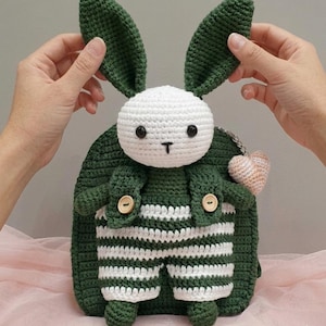 May include: A handmade crochet bunny backpack in green and white. The bunny has long green ears, a white head, and a black embroidered nose and eyes. The backpack features striped overalls and a small heart charm.