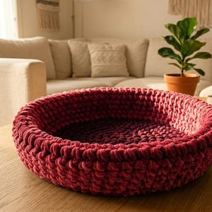 May include: A round, burgundy pet bed made of thick, textured yarn sits on a wooden coffee table. The bed is in a living room setting with a couch and potted plant in the background. The bed is approximately 20 inches in diameter.