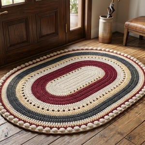Puede incluir: Una alfombra de ganchillo de forma ovalada con bandas concéntricas de hilo color crema, rojo oscuro, amarillo y gris oscuro. El borde exterior presenta un acabado decorativo de pequeñas borlas en relieve color crema con textura.