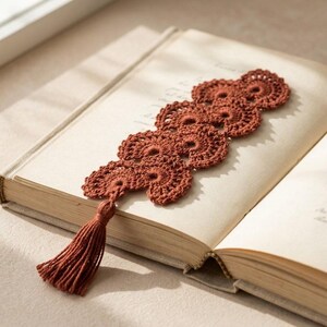 May include: A rust-coloured, handmade crochet bookmark with a tassel. The bookmark has a series of circular, openwork designs. It lies on the open pages of a vintage book, creating a cosy, literary aesthetic.