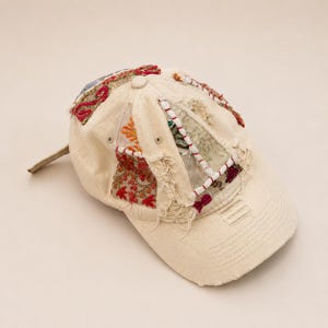 May include: A cream-coloured baseball cap featuring distressed fabric panels and intricate embroidery in shades of red, orange, and green. The cap includes frayed edges, visible stitching, and a curved brim with a worn, textured appearance.