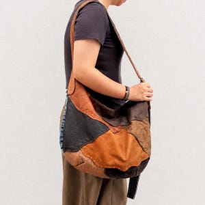 May include: A hobo-style shoulder bag constructed from patchwork leather panels in various shades of brown, tan, and dark charcoal grey. The bag features a long, integrated leather strap and a slouchy, unstructured silhouette with visible seams.