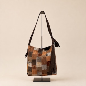 Può includere: Una borsa hobo con un design patchwork composto da vari pezzi di tessuto quadrati in tonalità marrone, beige, grigio e crema. La borsa presenta una lunga tracolla marrone scuro con un dettaglio a nodo su un lato e cuciture visibili.
