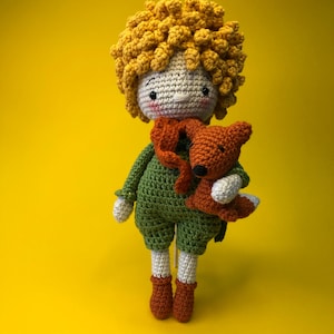 Peut inclure: Poupée au crochet faite à la main représentant un personnage aux cheveux bouclés jaunes, vêtu d'une tenue verte et tenant un petit renard orange. La poupée a un visage blanc avec des joues roses, sur fond jaune.