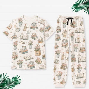 May include: Cream-colored pajama set with a short-sleeved top and pants. The set features a repeating pattern of books, flowers, teacups, and hearts. The pants have a black drawstring. The top has a crew neck.