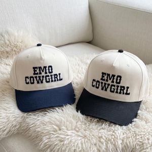 May include: Two baseball caps with the text "EMO COWGIRL" embroidered in black. The caps have a cream-colored crown and a navy blue and black brim. The caps are sitting on a white, fluffy surface.