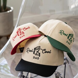 May include: Three beige baseball caps with the text "Reel Cool Dad" and a fish graphic. Each cap has a different colored brim: red, green, and black. The caps are displayed on a clear plastic chair.