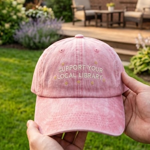May include: A pink baseball cap with a washed-out texture featuring the embroidered text SUPPORT YOUR LOCAL LIBRARY in white, accented by small yellow star icons surrounding the lettering on the front panel of the hat.