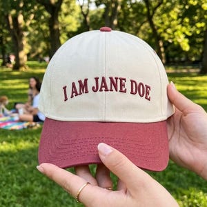 May include: A two-toned baseball cap featuring an off-white crown and a dark red brim. The front of the crown displays the embroidered text "I AM JANE DOE" in dark red capital letters. The cap has a small red button at the top center.