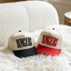 May include: Two baseball caps with the word "DINGERS" embroidered on the front. One cap has a black brim and black text, the other has a red brim and red text. Both caps are cream-colored.