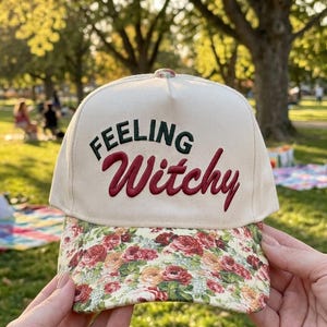 May include: A cream-colored baseball cap featuring the embroidered text "Feeling Witchy" in dark green and red. The brim of the cap is decorated with a colorful floral pattern consisting of red, orange, and yellow roses on a light background.