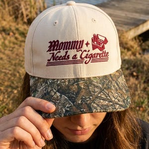 May include: A cream-colored baseball cap with a camouflage-patterned brim. The front features dark red embroidered text that reads Mommy Needs a Cigarette, accompanied by a small embroidered graphic of an open pack of cigarettes.