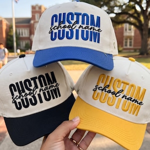 May include: Three baseball caps with off-white crowns and colored brims in blue, black, and yellow. Each cap features embroidered text reading CUSTOM school name CUSTOM in a bold, stacked font with a script font overlay in the center.