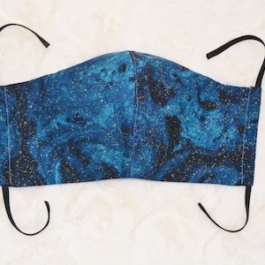K&ouml;nnte beinhalten: Eine blaue und goldene Gesichtsmaske mit Marmormuster. Die Maske hat schwarze elastische Ohrschlaufen.