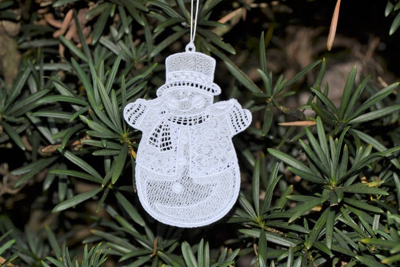 Pode incluir: Adorno de Natal de boneco de neve branco de croch&ecirc; com chap&eacute;u e cachecol. O adorno est&aacute; pendurado em um galho de &aacute;rvore.