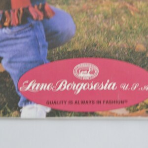 K&ouml;nnte beinhalten: Ein rotes ovales Etikett mit wei&szlig;em Text, der "Lamo Borgosesia U.P.A. QUALITY IS ALWAYS IN FASHION" lautet.