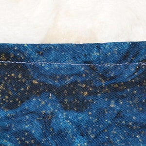 K&ouml;nnte beinhalten: Eine blaue Kordeltasche mit einem schwarzen Gummiband und einem goldenen Glitzermuster. Die Tasche ist mit einem hellblauen Faden gen&auml;ht.