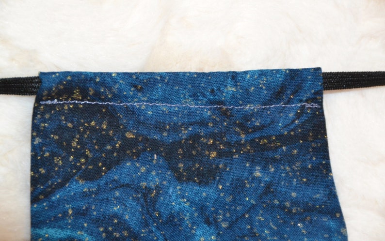 K&ouml;nnte beinhalten: Eine blaue Kordeltasche mit einem schwarzen Gummiband und einem goldenen Glitzermuster. Die Tasche ist mit einem hellblauen Faden gen&auml;ht.