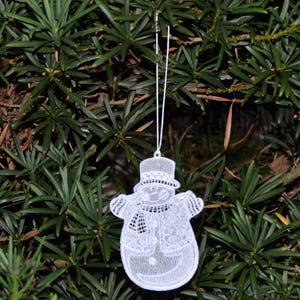 Pode incluir: Adorno de croch&ecirc; branco em forma de boneco de neve pendurado em um galho de uma &aacute;rvore de con&iacute;feras verde.