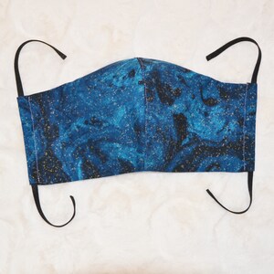 K&ouml;nnte beinhalten: Eine blaue und goldene Gesichtsmaske mit schwarzen elastischen Ohrschlaufen. Die Maske hat ein marmoriertes Muster mit goldenen Flocken.