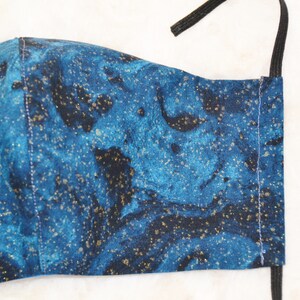 K&ouml;nnte beinhalten: Eine blaue und schwarze Gesichtsmaske mit goldenen Glitzer. Die Maske hat ein marmoriertes Muster und ist aus Stoff.