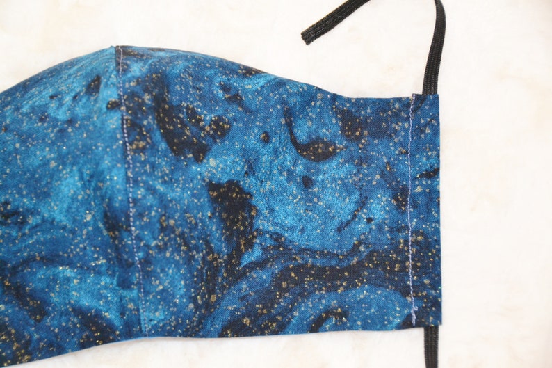 K&ouml;nnte beinhalten: Eine blaue und schwarze Gesichtsmaske mit goldenen Glitzer. Die Maske hat ein marmoriertes Muster und ist aus Stoff.