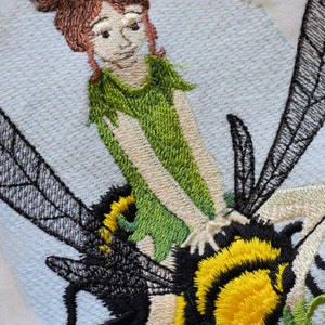 May include: Embroidered design of a fairy riding a bumblebee. The fairy has brown hair, a green dress, and is holding onto the bumblebee's back. The bumblebee is black and yellow with wings.