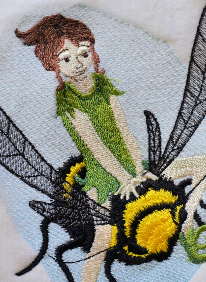May include: Embroidered design of a fairy riding a bumblebee. The fairy has brown hair, a green dress, and is holding onto the bumblebee's back. The bumblebee is black and yellow with wings.