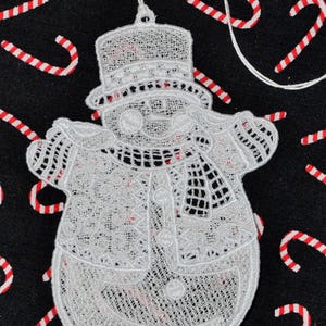 Pode incluir: Adorno de Natal de boneco de neve branco de croch&ecirc; em fundo preto. O boneco de neve tem um chap&eacute;u, uma echarpe e um bot&atilde;o.