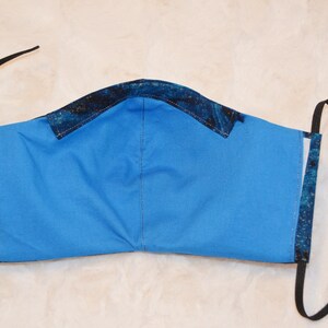 K&ouml;nnte beinhalten: Eine blaue Gesichtsmaske mit schwarzen verstellbaren B&auml;ndern. Die Maske besteht aus einem gemusterten Stoff mit blauem Hintergrund und schwarzen und goldenen Akzenten.