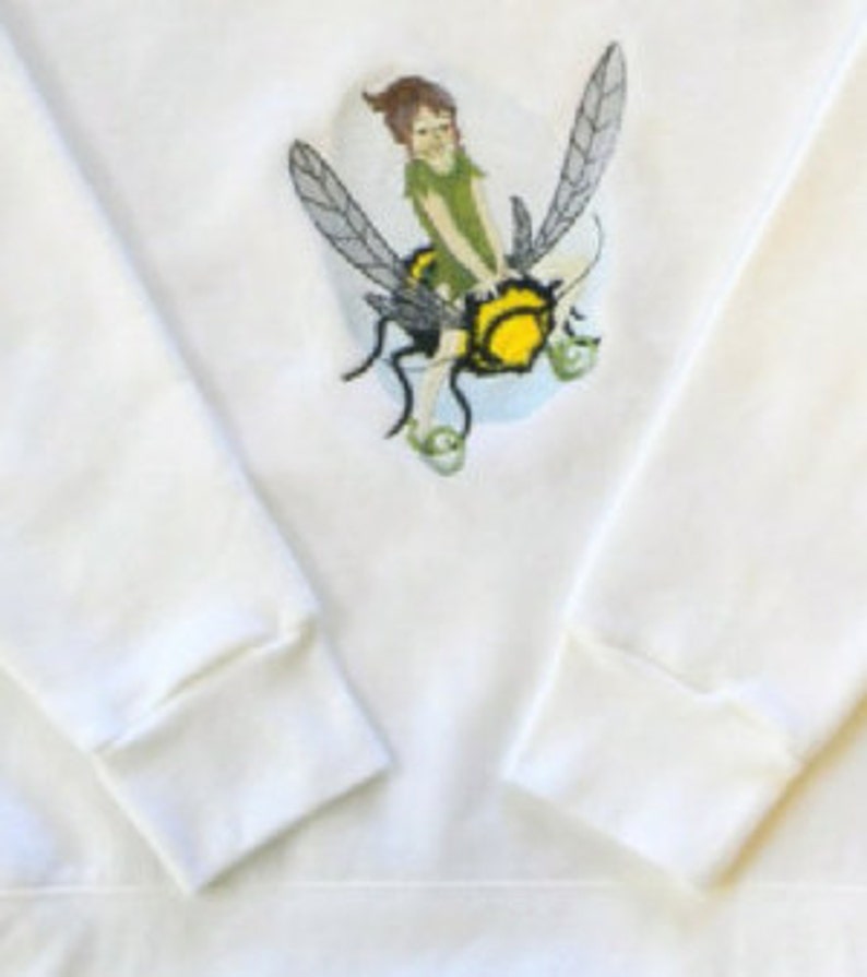 May include: White sweatshirt with a graphic of a fairy riding a bee. The fairy is wearing a green dress and has brown hair. The bee is yellow and black with wings.