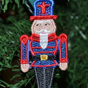 Puede incluir: Un adorno de cascanueces bordado en rojo, azul y blanco con un sombrero negro y una barba blanca. El adorno está colgado de un árbol de Navidad verde.