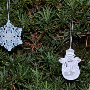 Pode incluir: Dois enfeites de Natal de renda branca pendurados em uma &aacute;rvore verde. Um enfeite &eacute; um floco de neve e o outro &eacute; um boneco de neve.
