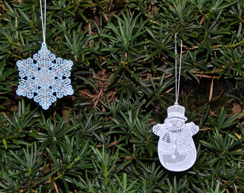 Pode incluir: Dois enfeites de Natal de renda branca pendurados em uma &aacute;rvore verde. Um enfeite &eacute; um floco de neve e o outro &eacute; um boneco de neve.