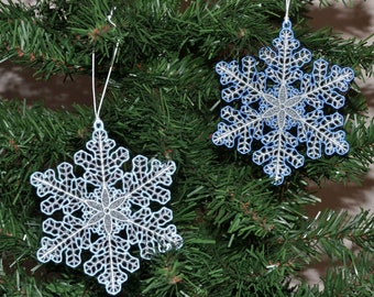 Pode incluir: Dois enfeites de floco de neve branco com detalhes azuis pendurados em uma árvore de Natal verde.