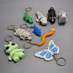 May include: A collection of novelty keychains featuring various animal and creature designs. Includes a green alligator, a white shark, a black hippo, a white hippo, a gray mouse, a blue dolphin, an orange worm, a green alien, and a butterfly with blue accents.