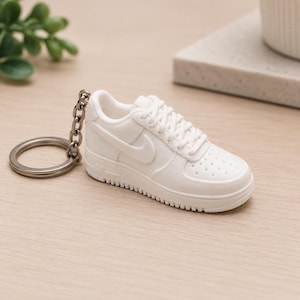 Op de afbeelding: Een witte miniatuur sneaker sleutelhanger met een zilveren sleutelring. De sneaker heeft het Nike swoosh logo en een textuurzool. De sleutelhanger is een kleine, gedetailleerde replica van een klassieke sportschoen.
