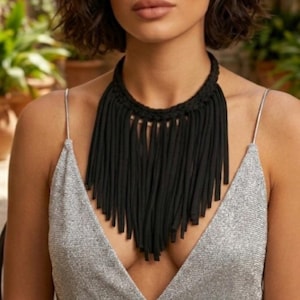 May include: A black necklace featuring a thick braided collar and long, vertical fringe strands that hang down the chest. The necklace is worn with a silver-coloured sleeveless top that has thin spaghetti straps and a deep V-neckline.