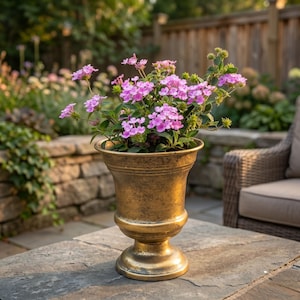 May include: A bronze-colored urn-shaped planter filled with vibrant pink flowers. The planter has a pedestal base and is sitting on a stone surface. The background includes a stone wall, wooden fence, and outdoor furniture.