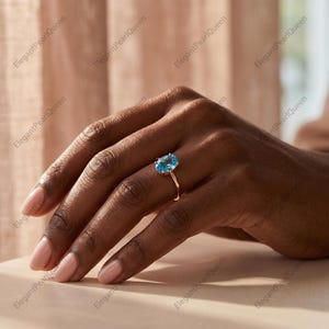 Peut inclure: Une bague ornée d'une pierre précieuse bleue taille ovale montée sur un anneau fin en or poli. La pierre est d'un bleu ciel vif et clair, maintenue par un sertissage à quatre griffes qui surélève la gemme au-dessus de l'anneau métallique.