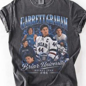 以下が含まれることがあります： ホッケー選手のグラフィックが特徴のチャコールグレーのTシャツ。テキストには「GARRETT GRAHAM」と「Briar University Hockey Team #44」が含まれています。デザインには、ユニフォームを着たホッケー選手の画像とBriar Universityのロゴが含まれています。