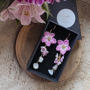 May include: Handmade pink flower earrings with gold-tone hooks and white pearl details, presented in a black box. The earrings are part of the "Malik Handmade" collection. The background features pink flowers and a wooden surface.